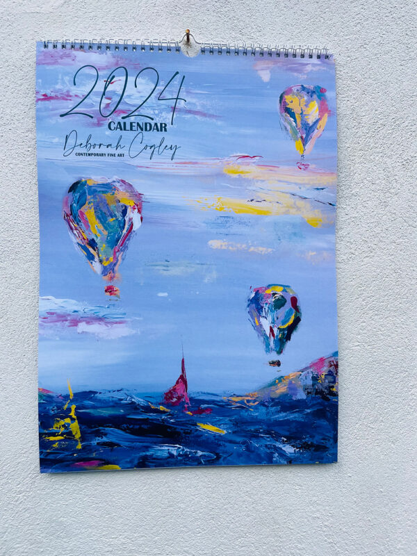 Front of calendar depicting air balloons and boats on the sea with a mix of blues, pinks and yellows